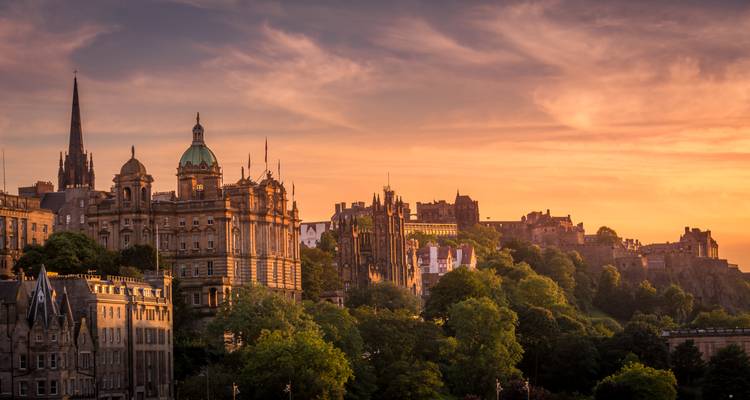 Edinburgh's historic skyline glows under a dramatic sunset with the castle and spires rising above lush trees.