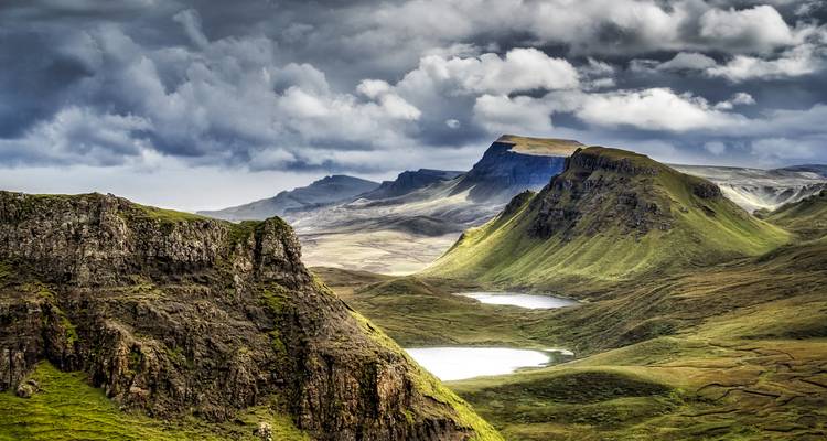 Dramatic, rugged green ridges and cliffs of the Isle of Skye sweep toward distant lochs under a moody sky.