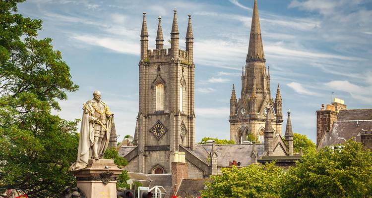 Neo-Gothic church spires and a statue rising above leafy trees in a Scottish cityscape