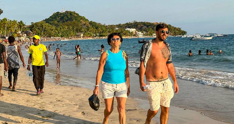 Personas caminando por una playa con barcos en el agua.