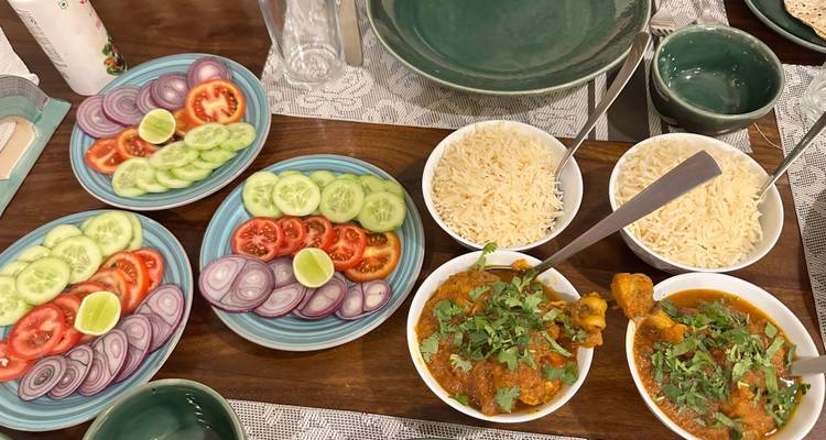 Indische hausgemachte Mahlzeit mit Schüsseln voller Curry, Basmatireis und ordentlich angerichteten Tellern mit geschnittenen Tomaten, Zwiebeln, Gurken und Limetten auf einem Holztisch.