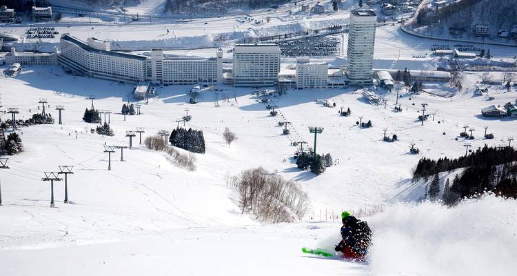 Ein Skifahrer fährt eine breite Alpinhang hinab und überblickt einen ausgedehnten Skiresortkomplex, der in einem verschneiten Tal eingebettet ist.