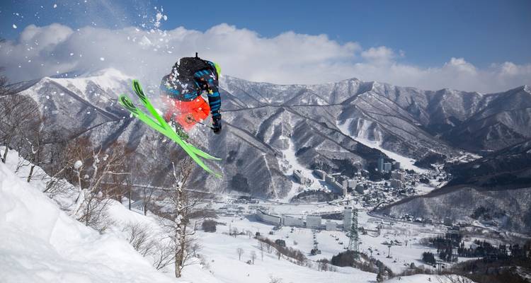 Ein Skifahrer in leuchtend farbiger Ausruestung fuehrt einen Sprung abseits der Piste durch und ueberblickt ein schneebedecktes Bergtal und das Dorf darunter unter einem klaren blauen Himmel.