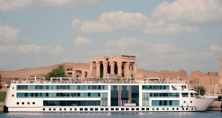 Navire de croisière fluviale moderne aux parois vitrées passant devant les ruines antiques du temple de Kom Ombo le long du Nil.