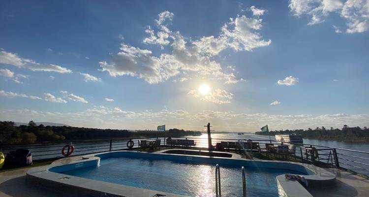 Terrasse de piscine ensoleillée sur le toit d'un bateau de croisière sur le Nil avec paysage fluvial sous un ciel lumineux.