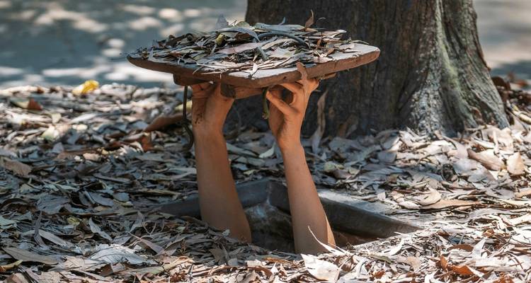Handen die tevoorschijn komen uit een gecamoufleerd tunneldeksel bedekt met bladeren, onderdeel van de Cu Chi Tunnels.