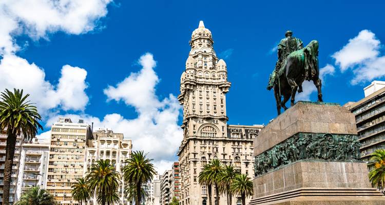 Blue-sky view of Palacio Salvo and Artigas statue framed by palm trees on Plaza Independencia