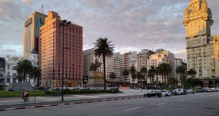 View of Plaza Independencia in Montevideo with palm trees, historic and modern buildings under warm evening light, a couple walking in the foreground