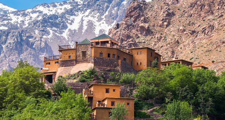 Una casa de huéspedes en tonos tierra se aferra a una ladera con montañas nevadas de fondo.