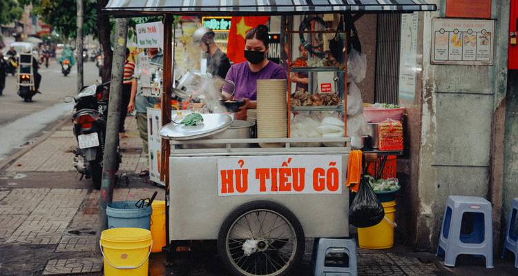 Street-food vendor prepares dishes at a small cart decorated with Vietnamese signage on a city sidewalk.