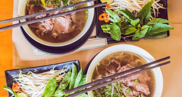 Top-down view of two steaming bowls of Vietnamese pho with fresh herbs, sprouts and chopsticks laid out on a table.