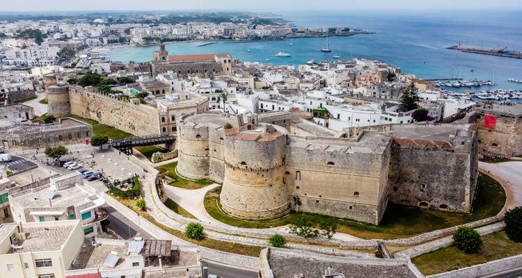 Ville fortifiée côtière dominant une marina et la mer ouverte sous un ciel dégagé.