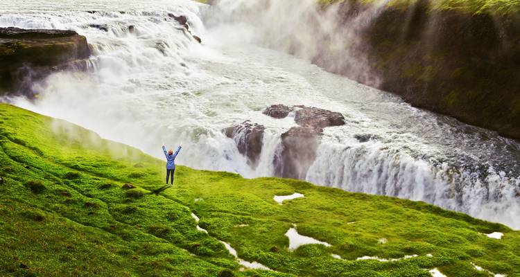Een solo reiziger die juicht naast de machtige watervallen en nevel van de Gullfoss waterval.