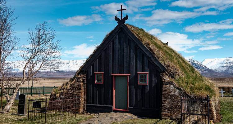 Traditionele IJslandse plaggen-daken kerk met zwarte houten gevel tegen sneeuwbedekte bergtoppen.