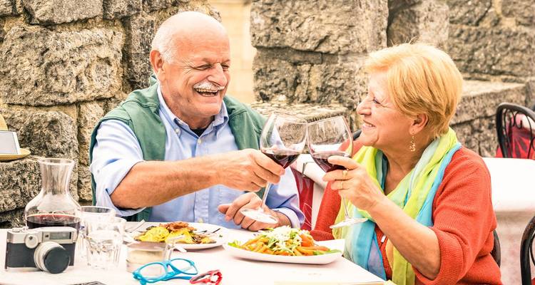 Pareja mayor sonriendo brindando con copas de vino mientras disfrutan de una comida italiana al aire libre.