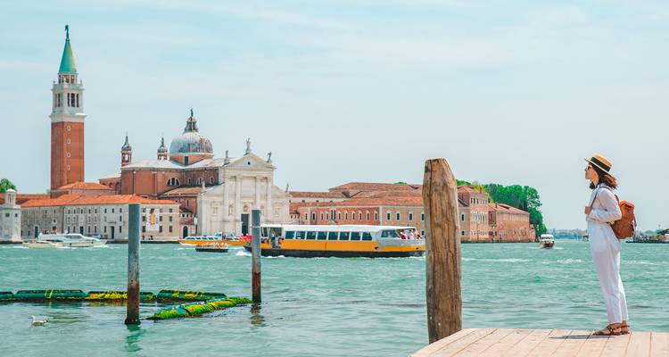 Vrouw die op een pier staat en uitkijkt over de skyline van Venetië terwijl er een boot voorbijvaart.