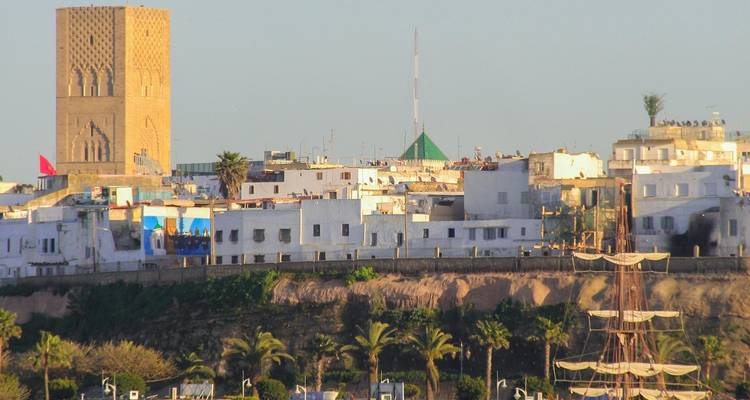 View of white hillside buildings and the sandstone Hassan Tower with greenery and palm trees in front.