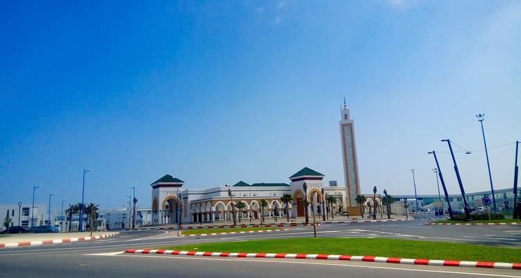 Wide boulevard roundabout leading to a white-and-tan building with Islamic arches and a tall slender minaret under blue sky.