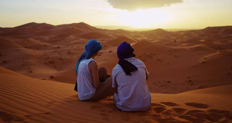 Zwei Reisende mit Turbanen sitzen auf einer Sanddüne und beobachten den Sonnenuntergang über der welligen Sahara-Wüste.