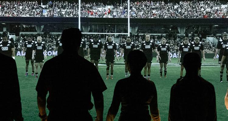 Espectadores en silueta observan a jugadores de rugby realizar la haka en un campo de estadio brillantemente iluminado por la noche.