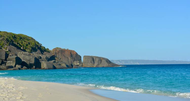 Plage de sable avec une eau bleue claire et des falaises rocheuses.