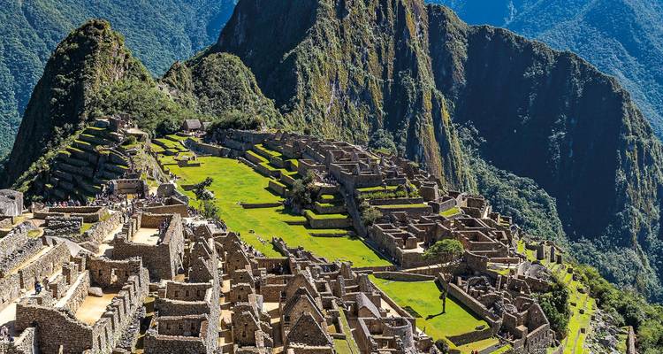 Ein weiter Blick auf die antiken Steinterrassen und Tempel von Machu Picchu, eingerahmt von üppigen Andengipfeln unter strahlendem Sonnenschein.