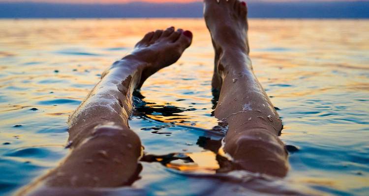 Person, die bei Sonnenuntergang auf dem Rücken im Toten Meer treibt, mit Beinen und Füßen über dem Wasser