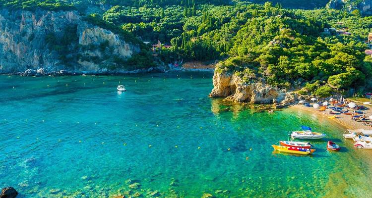 Kristalhelder aquamarijnblauw baaitje met kleine bootjes en beboste kliffen langs de kust van Corfu.