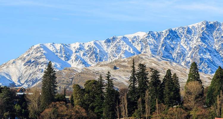 Cordillera montañosa cubierta de nieve que se eleva sobre pinos verde oscuro y un pequeño asentamiento bajo un cielo azul claro cerca de Queenstown.
