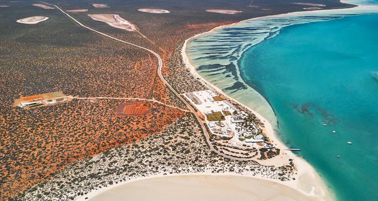 Vue aérienne d'une baie turquoise rencontrant un désert de couleur rouille, avec un complexe de villégiature de faible hauteur sur le littoral.