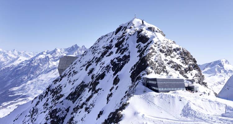 Ein schneebedeckter Alpengipfel mit einer modernen Beobachtungsstation und dramatischer Bergkulisse unter einem klaren blauen Himmel.