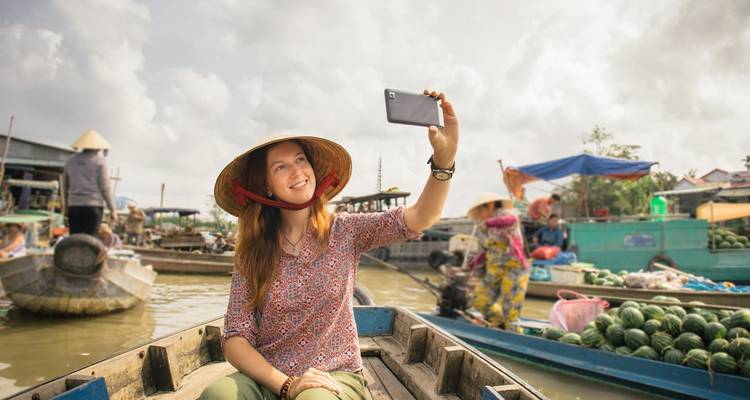 Female traveller taking selfie from a wooden boat amid floating market activity.