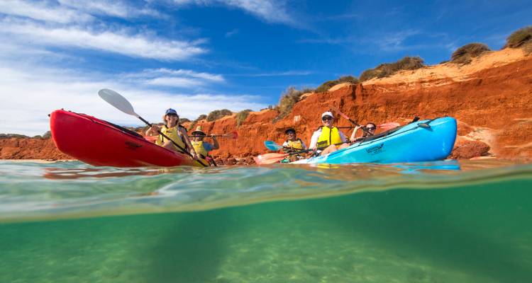 Group of kayakers paddling near red coastal cliffs, half-submerged camera showing clear green water
