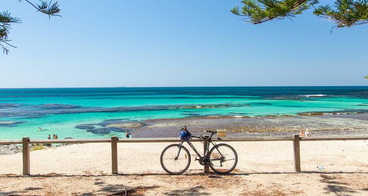 Wide sandy beach with turquoise water; a bicycle leans on a simple fence while swimmers enjoy the shallows