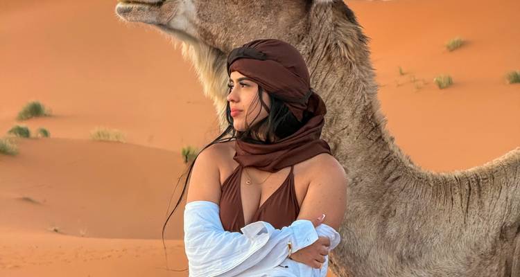 Jeune femme portant un foulard posant à côté d'un chameau face aux dunes de sable orange ondulantes.