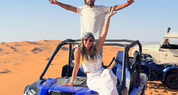 Un couple souriant pose sur un buggy tout-terrain bleu au milieu des dunes ondulantes orange du désert du Sahara.