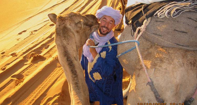 Hombre vestido con ropa bereber azul interactuando de manera divertida con un camello en las dunas del desierto iluminadas por el sol (marca de fecha visible)