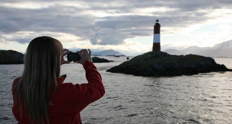 Ein Reisender in einer roten Jacke fotografiert einen einsamen Leuchtturm auf einem felsigen Eiland in einem dramatischen patagonischen Kanal.