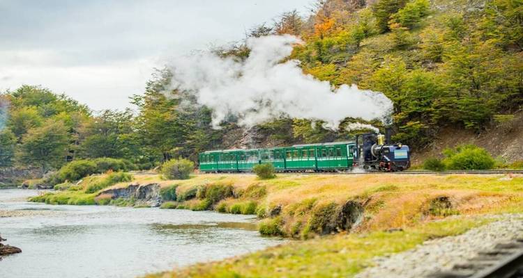 Ein vintage grüner Dampfzug fährt neben einem gewundenen Fluss durch einen farbenfrohen Herbstwald in Tierra del Fuego.