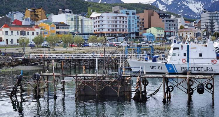 Farbenfrohe Hafengebäude und ein Patrouillenboot säumen den Hafen von Ushuaia mit schneebedeckten Gipfeln im Hintergrund.