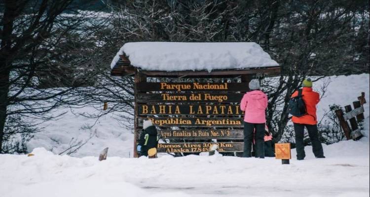 Gebündelte Wanderer lesen ein schneebedecktes Schild des Tierra del Fuego Nationalparks in einer Winterlandschaft.