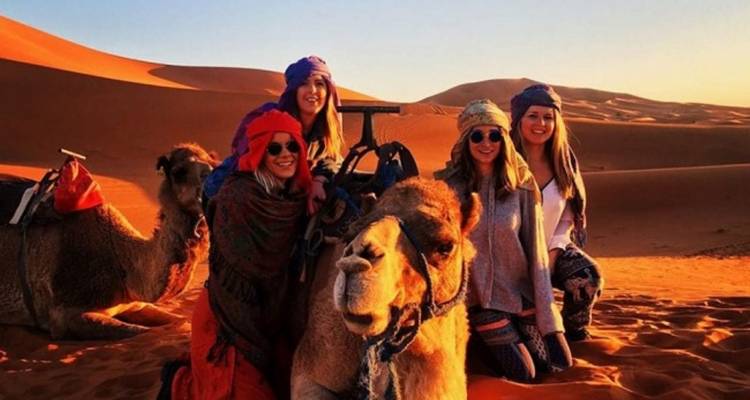 Gruppe von Menschen mit Kamelen in einer Wüstenlandschaft bei Sonnenuntergang.