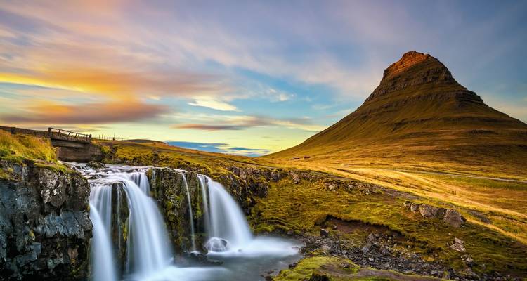 Picturesque pointed mountain beside tiered waterfall at sunset with colorful sky.