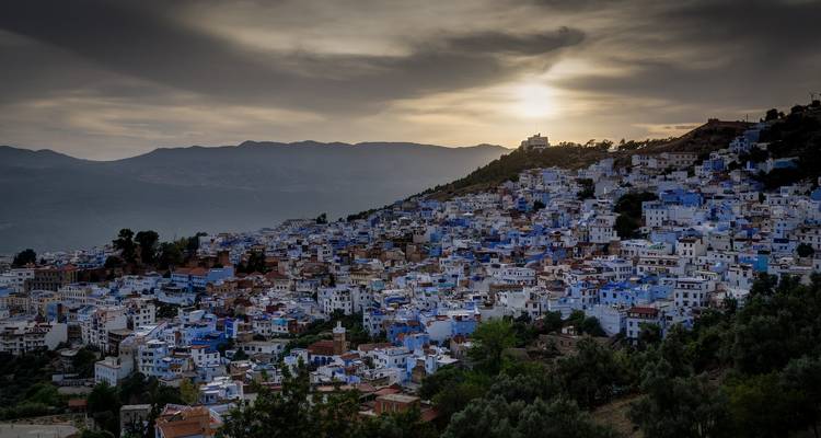 Chefchaouen strekt zich uit over een heuvelflank in de schemering met blauwe huizen en verre bergen onder een gloeiende hemel