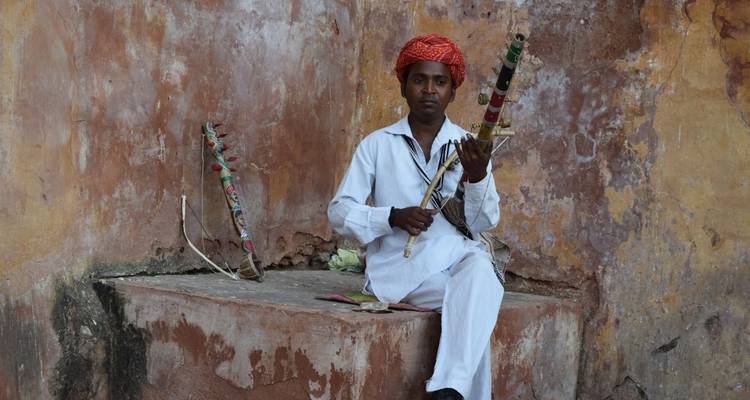 A man seated playing a string instrument against a rustic background.