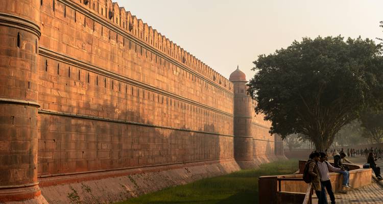 Red fort wall with trees and people walking nearby.