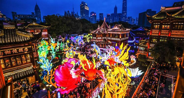 Levendige lantaarnfestival in Shanghais Yuyuan Garden met kleurrijke verlichte draak- en visfiguren te midden van traditionele gebouwen en de skyline van de stad op de achtergrond.