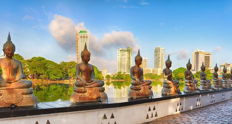Boeddhabeelden langs een stadswaterkant, met moderne wolkenkrabbers op de achtergrond.