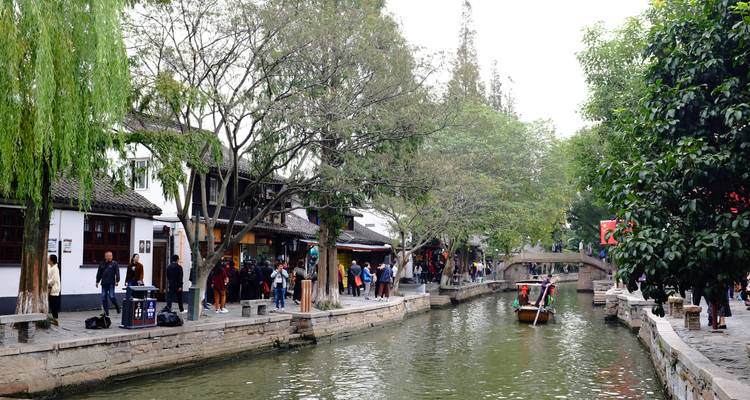 Calle junto al canal del pueblo acuático de Zhujiajiao con turistas caminando y una barca de canal pasando bajo un puente de piedra.