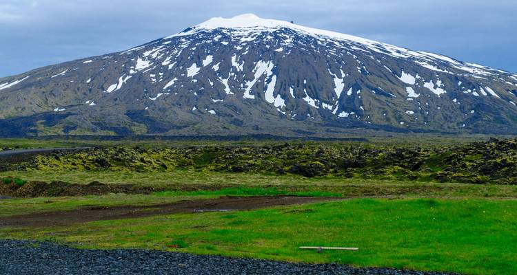 Stratovolcan aux sommets enneigés s'élevant au-dessus de champs de lave recouverts de mousse sous un ciel nuageux dans l'ouest de l'Islande.